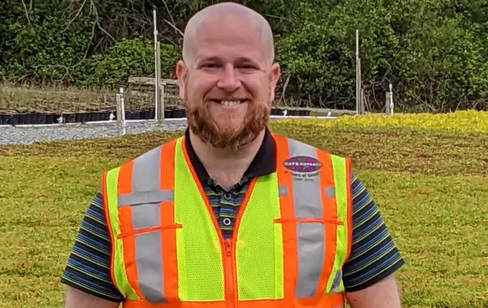 An image of a man standing on a roof with vegetation in the background. He is smiling and wearing a bright hi-visibility vest.